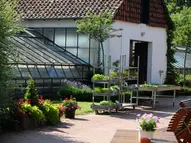 Gewächshaus und historisches Gebäude, weiß mit dunklem Dach. Auf dem Vorplatz stehen Blumen und Pflanzen